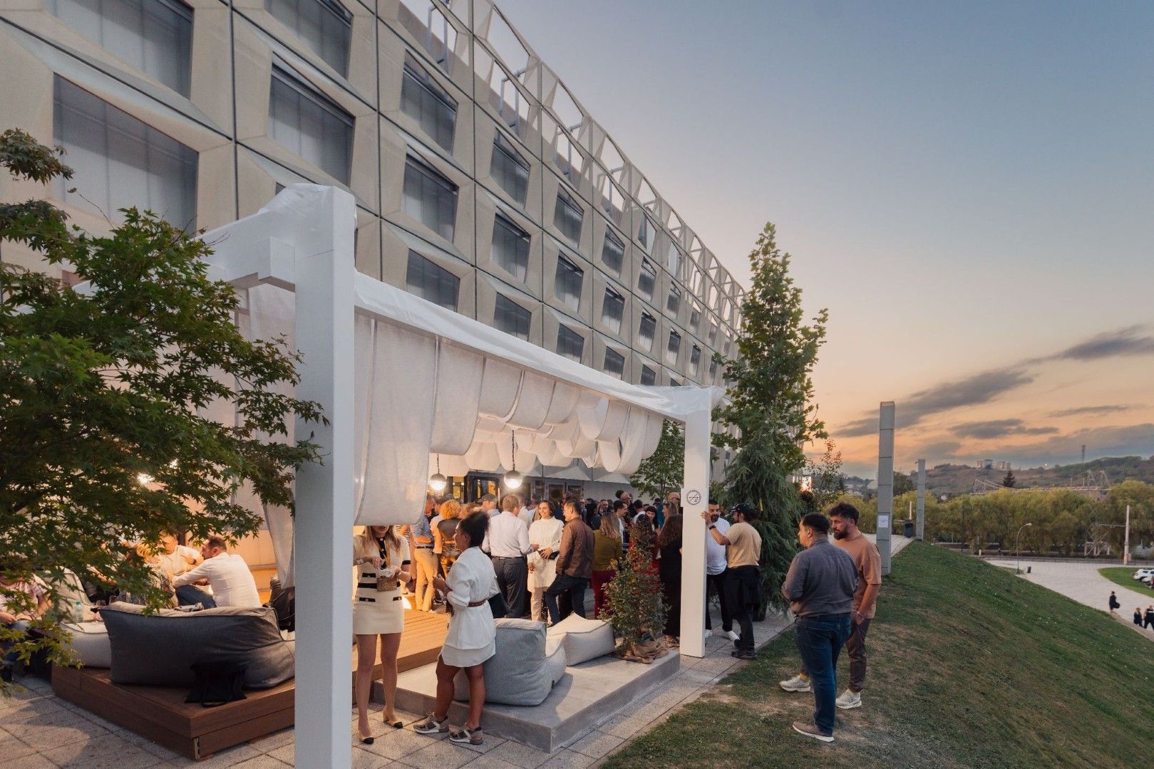 Sunlit — pergolă albă cu perdele textile și podium din lemn, în fața unei fațade cu panouri prefabricate
