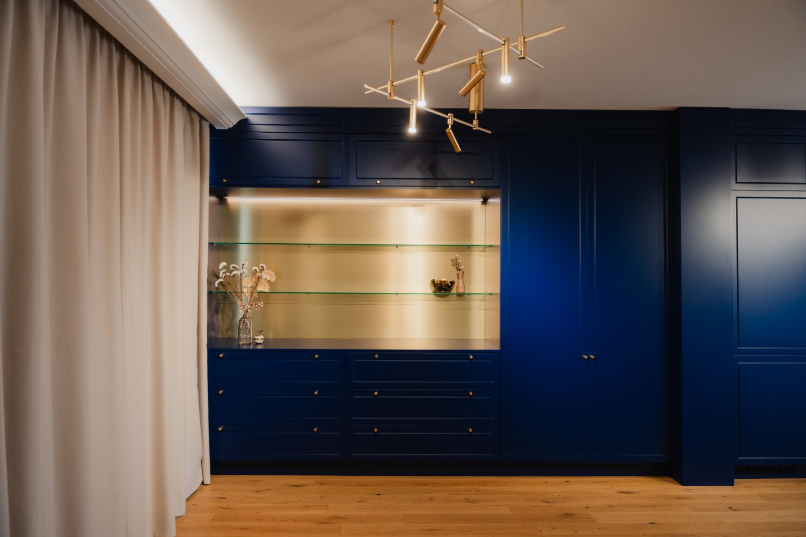 Office — perete de mobilier albastru RAL 5013 cobalt blue, cu rafturi din sticlă pe fundal metalic auriu și pendul cu brațe metalice