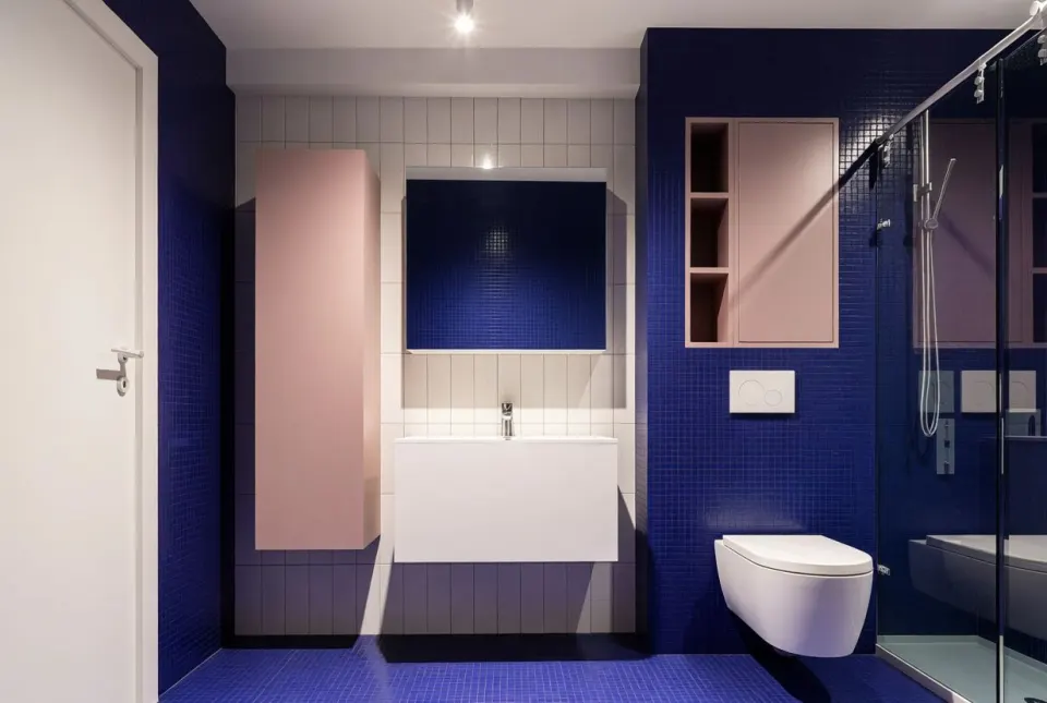 Apartament AD 1 — baie cu mozaic albastru, corpuri roz suspendate, vas WC suspendat și cabină de duș cu sticlă