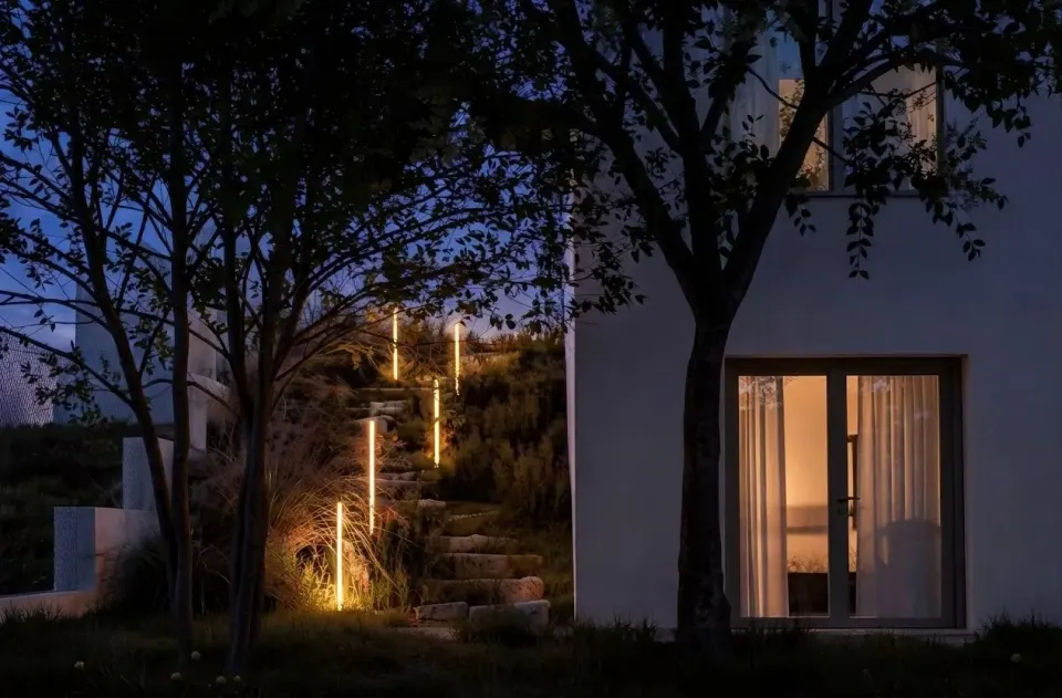 Casa MV — ușa-fereastră a parterului iluminată noaptea și scara exterioară pe pantă, marcată de stâlpi luminoși
